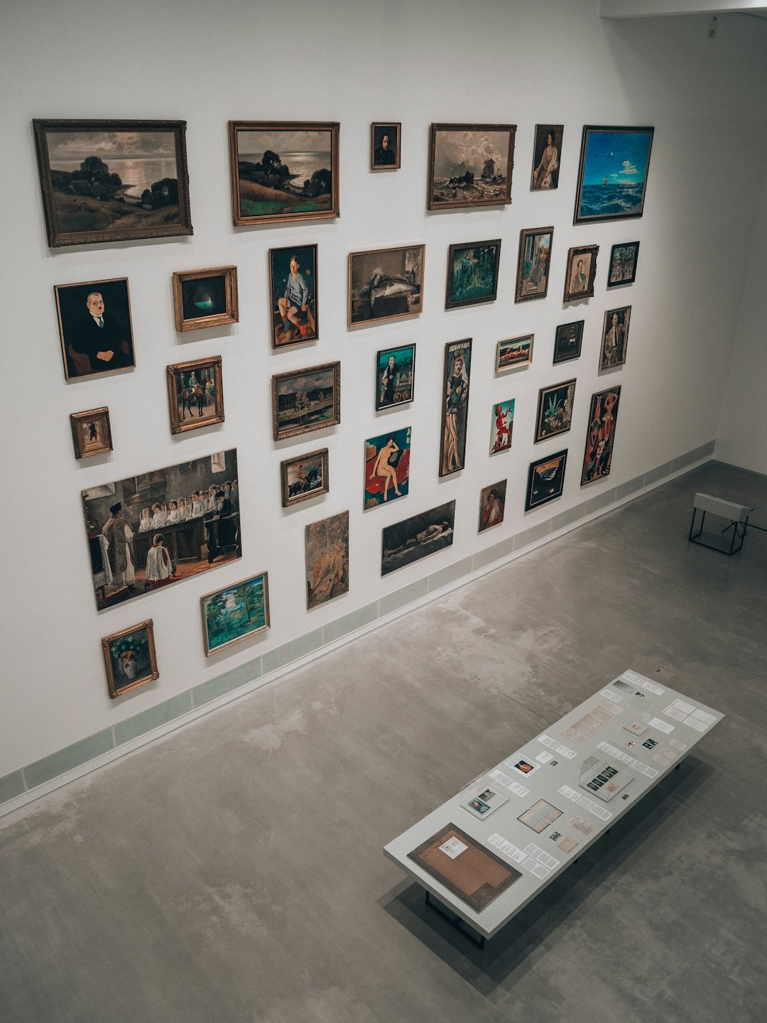 Gerahmte Gemälde hängen über Broschüren auf einem Ausstellungstisch in einer Galerie.